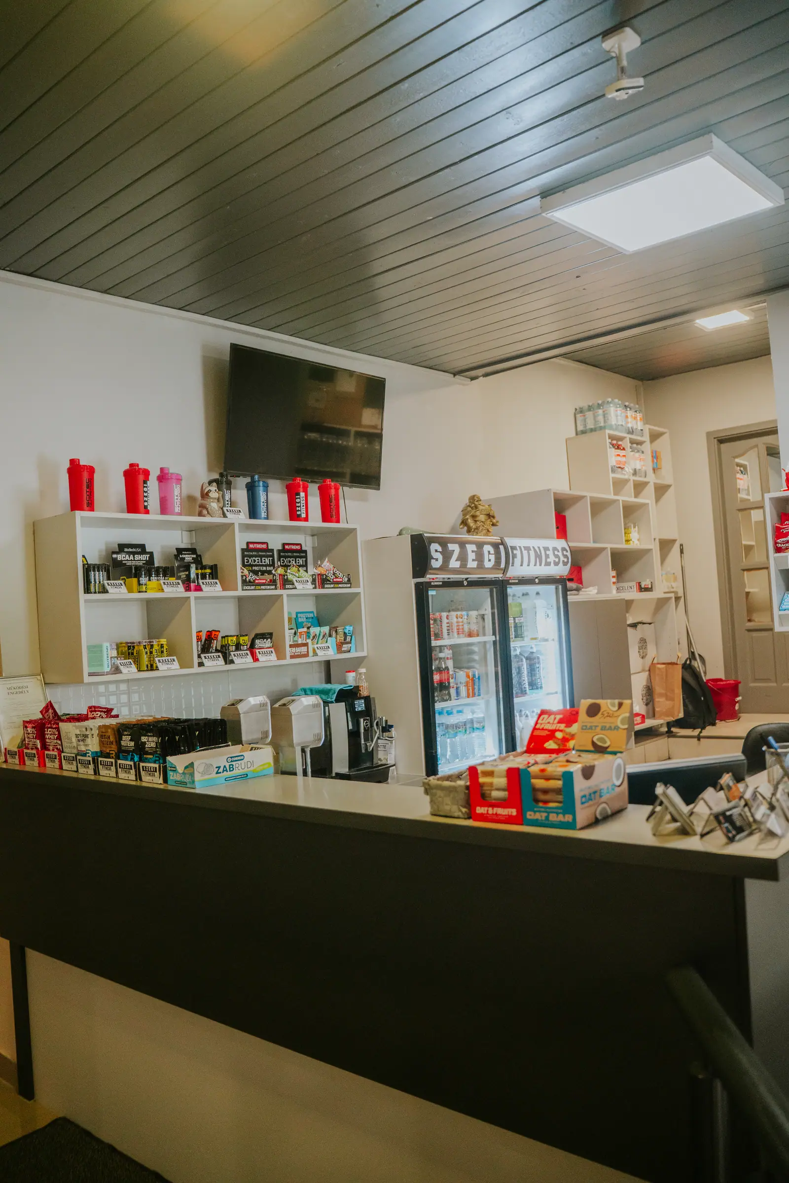 Recepciós pult a shop polcaival, a falon tévé, háttérben a Szegi Fitness hűtő